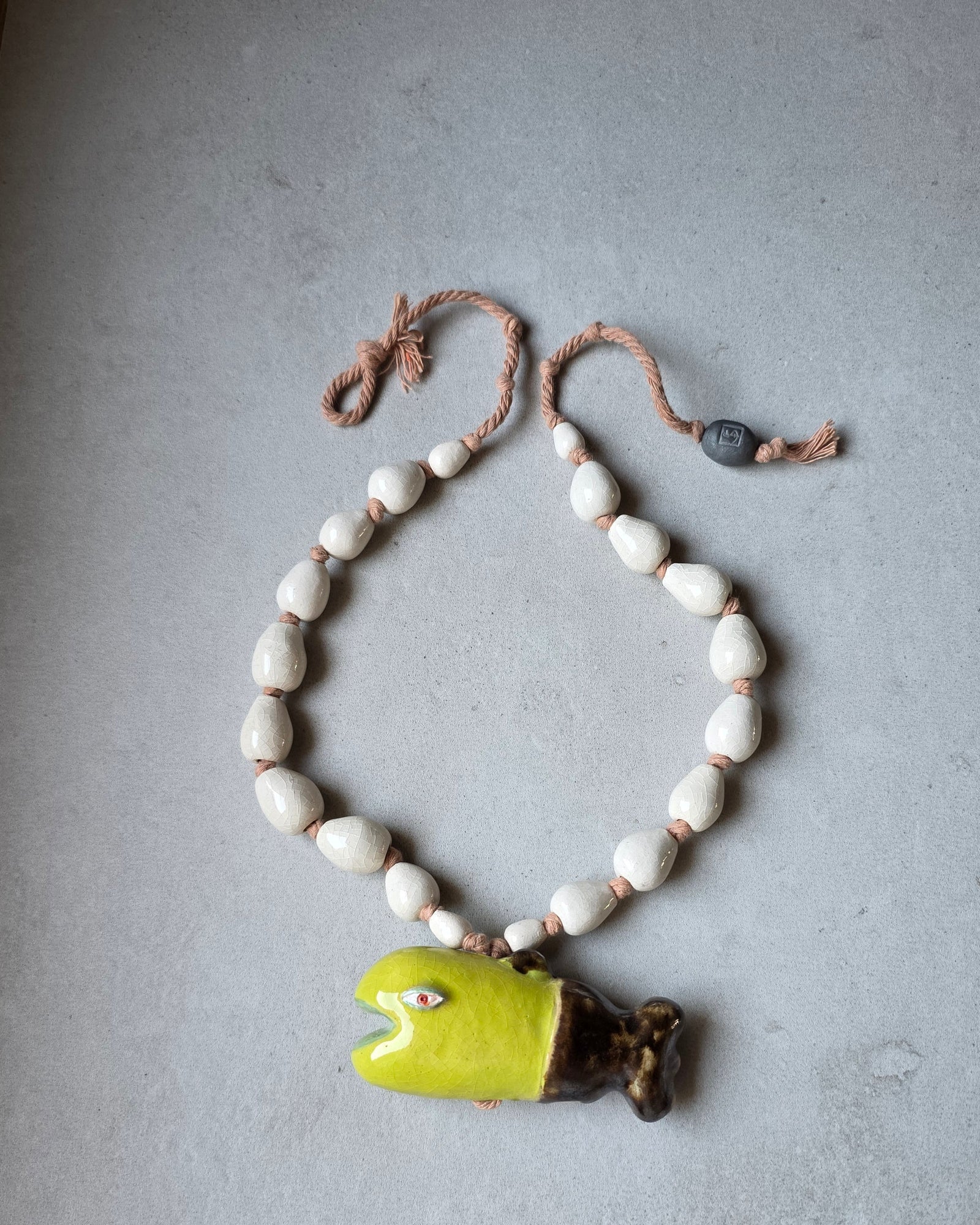 Collana Pesce Alga – distesa ad anello su fondo grigio, pesce verde lime con coda bronzo, perle a goccia bianco avorio su corda di cotone rosata e perla grigia con logo SF