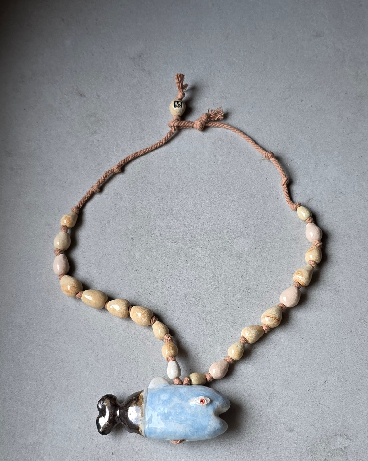 Collana Pesce Azzurro – distesa ad anello su fondo grigio, pesce azzurro con coda scura, perle a goccia sabbia e rosa cipria su corda di cotone e perla avorio con logo SF