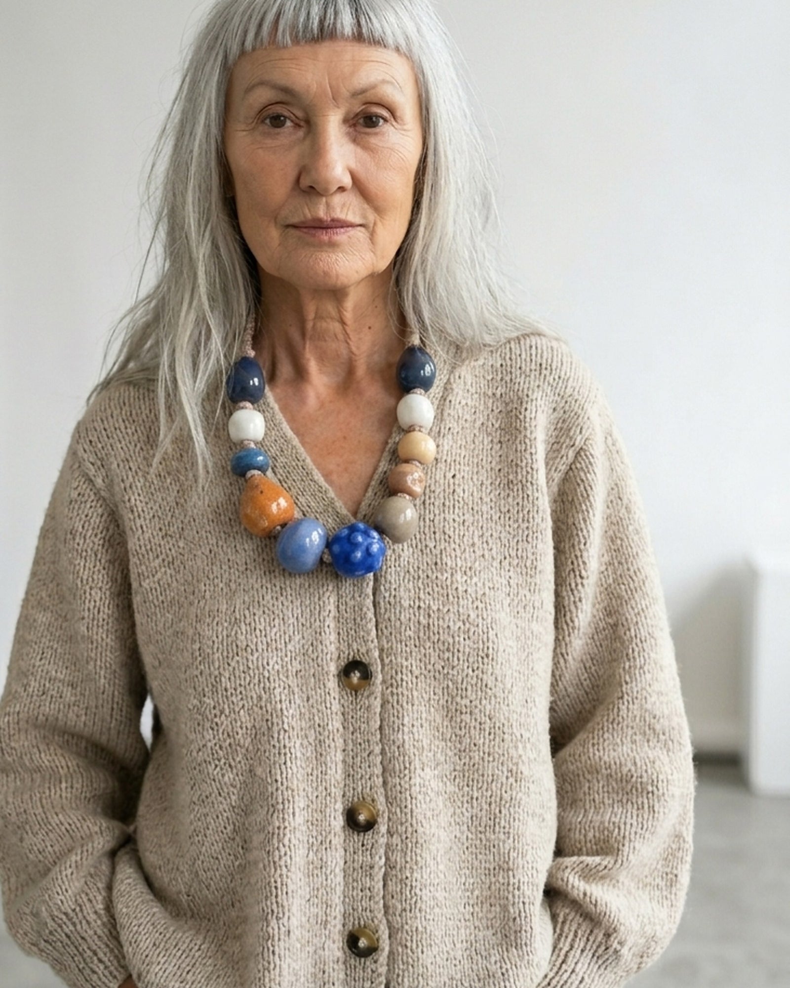 Donna con capelli grigi e cardigan beige indossa la collana Mediterraneo, grandi perle in ceramica blu, bianche, ocra e arancio su cordino naturale. Il volto è immaginato, il gioiello è vero: fatto a mano in atelier.