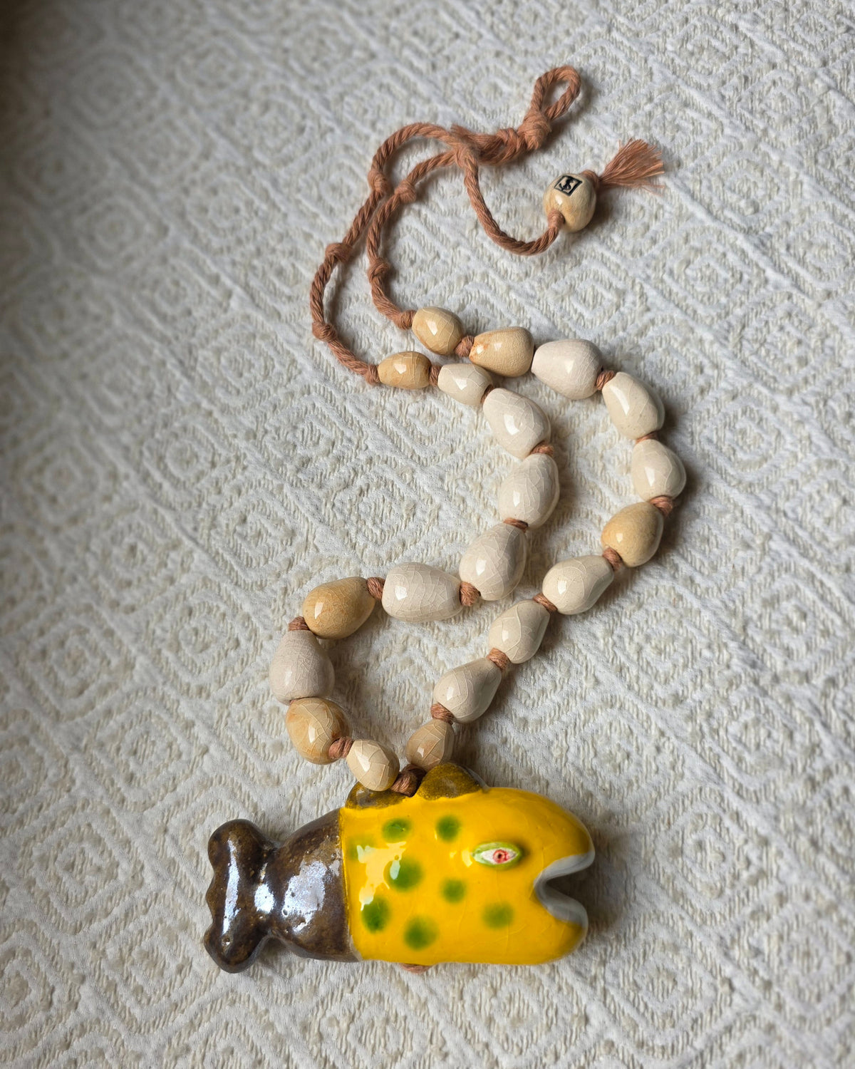 Collana Pesce Raro – distesa su un tessuto a rilievo, pesce giallo a pois verdi con coda bronzo, perle a goccia bianco avorio e sabbia su corda di cotone rosata