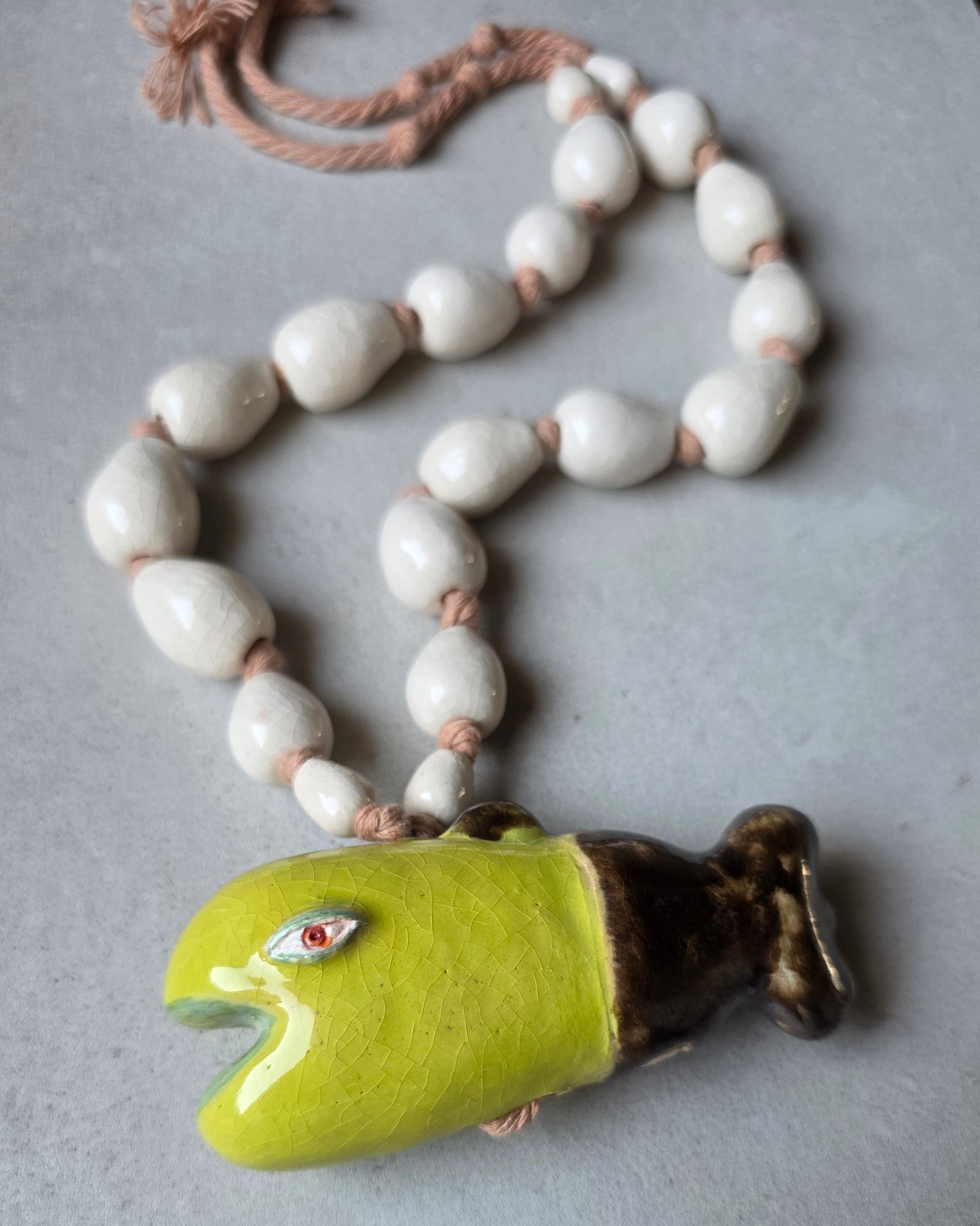 Collana Pesce Alga – distesa ad anello su fondo grigio, pesce verde lime con coda bronzo, perle a goccia bianco avorio su corda di cotone rosata e perla grigia con logo SF