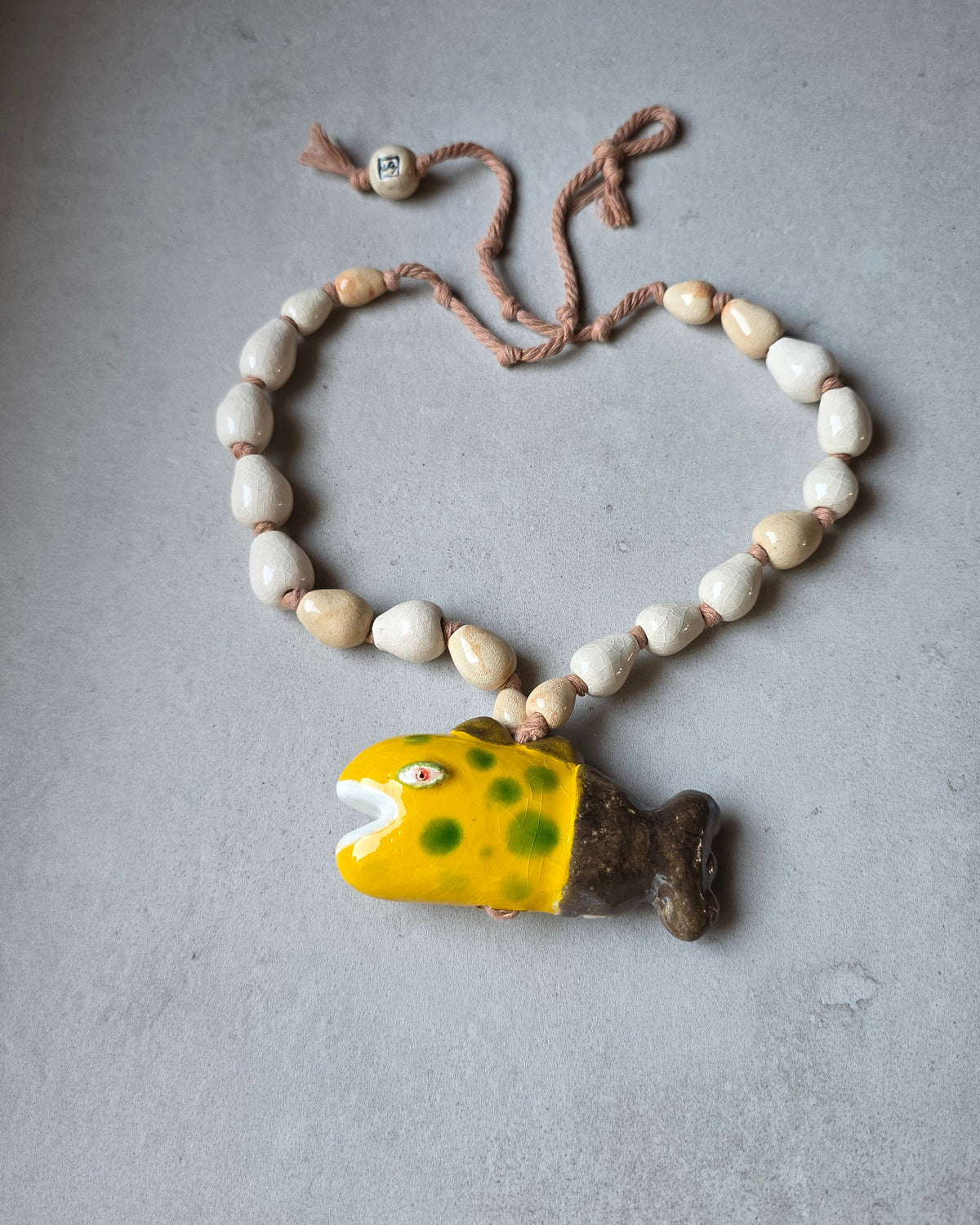 Collana Pesce Raro – disposta a cuore su fondo grigio, con il pesce giallo in basso e le perle a goccia craquelé