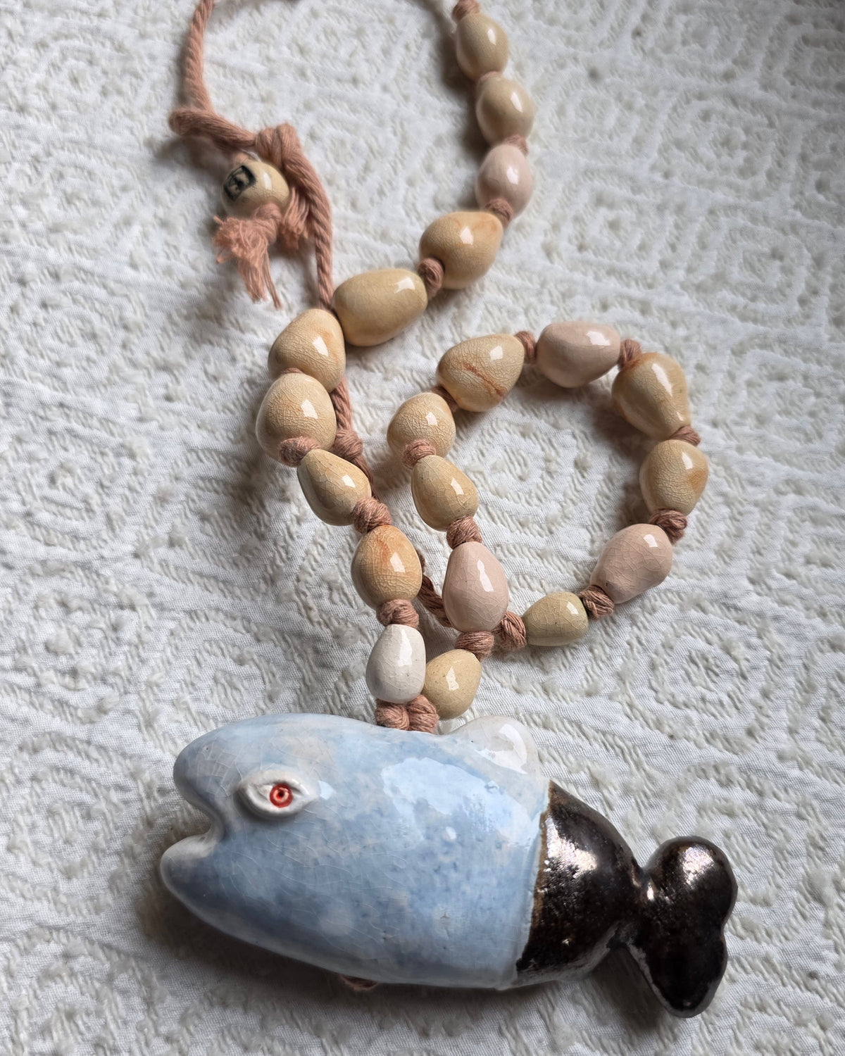 Collana Pesce Azzurro – la collana distesa su un tessuto crema, il pesce azzurro con la coda scura sotto le perle sabbia e rosa, la perla avorio SF sulla corda