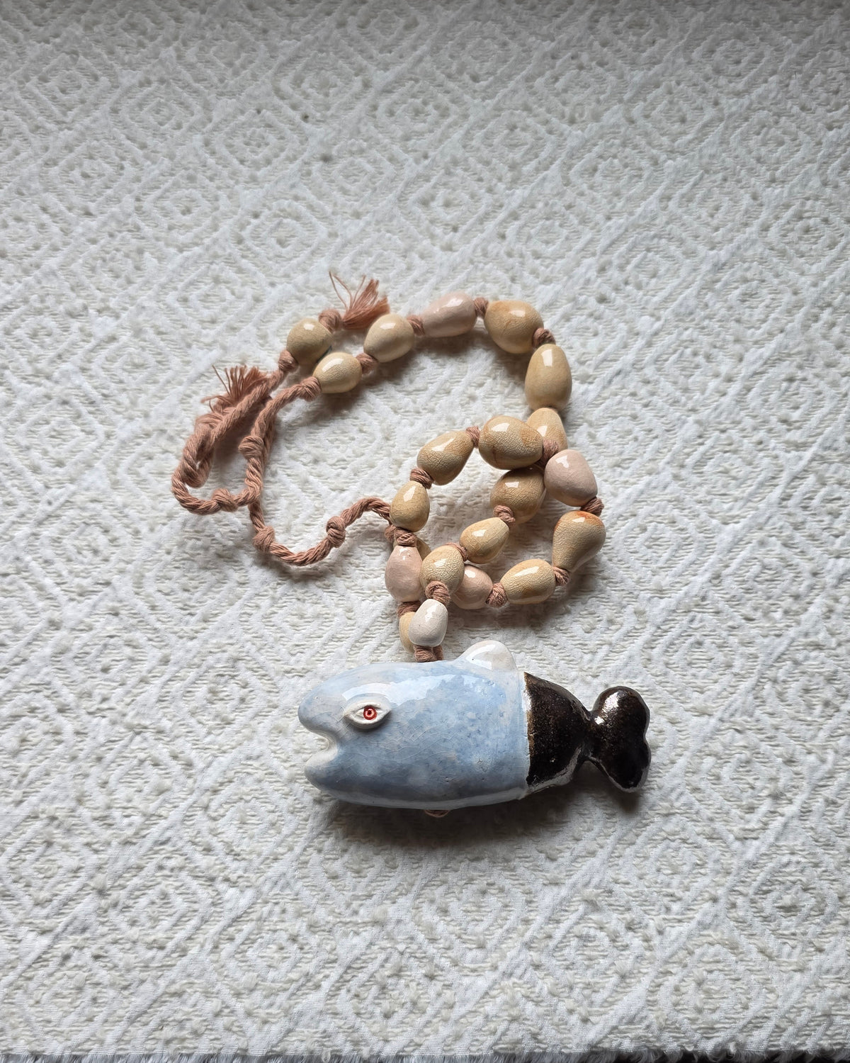 Collana Pesce Azzurro – la collana vista dall'alto su un tessuto crema, il pesce azzurro sotto un morbido cappio di perle sabbia e rosa